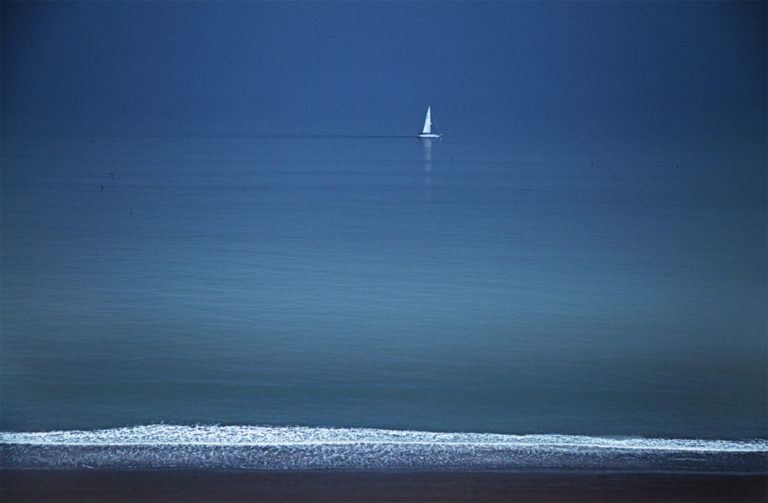 A colour photograph of a sailing boat on a calm sea at dusk passing by Cromer Norfolk.