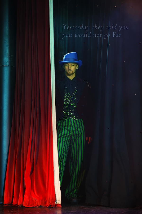 A dancer stares out from behind a stage curtain. He's looking thoughtfully at the audience. A Theatrical quotation makes sense of the scene.