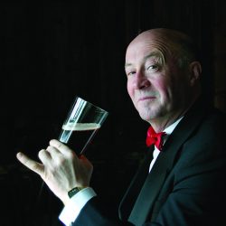 A man in a dinner jacket and red bow tie holds a pint glass of beer with a discerning look.